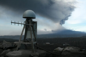 Instrumentation on the top of a mountain with a volcano erupting in the background and plumes of dark clouds