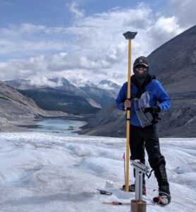 man standing on ice with testing equipment, mounts in the background