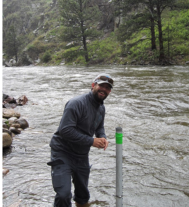 Man standing in a fast river smiling while testing the water with a long gray tube