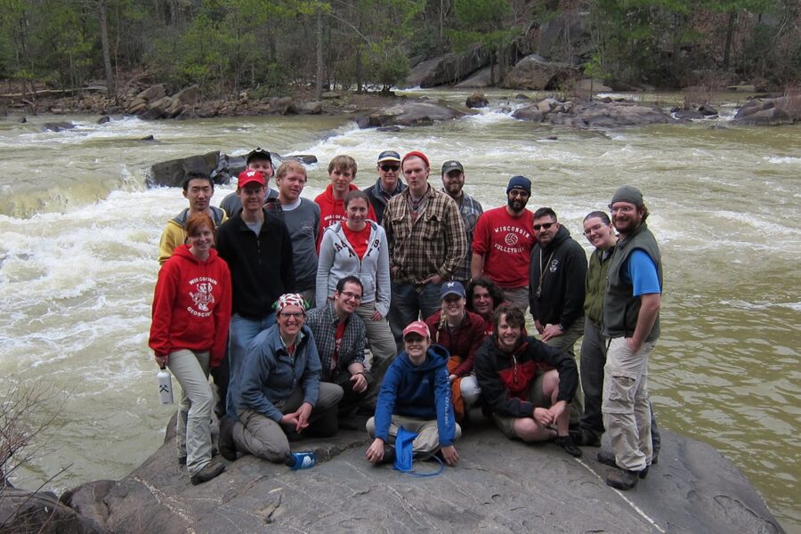 A group stands on a big rock, a rushing river behind them.