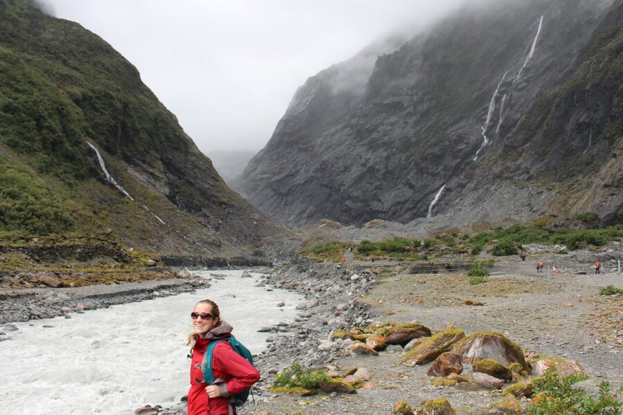 Ann Bauer standing next to a rushing stream with foggy mountains and waterfalls in the background.