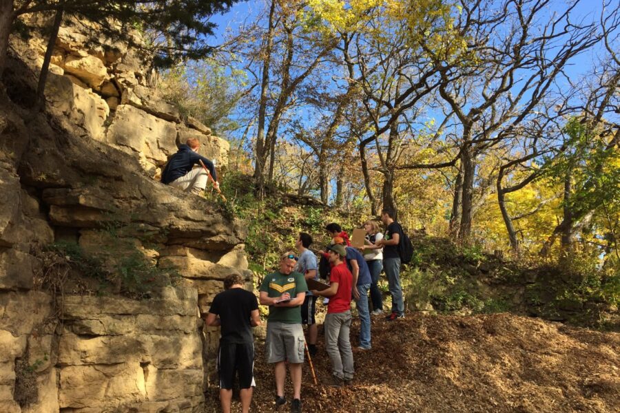Geoscience students study and take notes next to a rock formation in a wooded area in autumn.