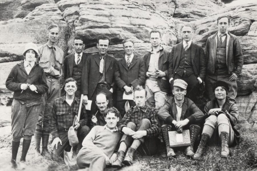 About a dozen men and women sit and stand in front of a rock formation on a field trip for a Glacial Geology class